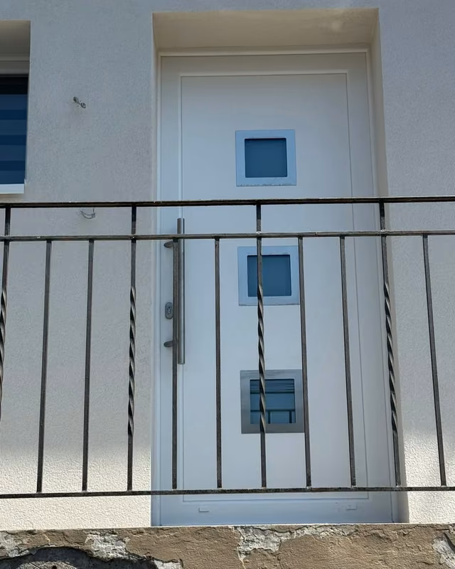 Porte d’entrée blanche à trois petites fenêtres carrées et poignée verticale, derrière un garde-corps — photo de chantier